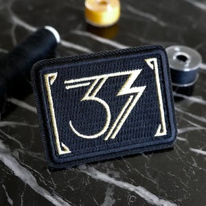 Peut inclure: Écusson rectangulaire bleu marine brodé du chiffre "33" en or, de style art déco. L'écusson a une bordure noire. Des bobines de fil et un petit objet jaune sont en arrière-plan.