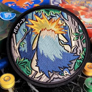 Peut inclure: Écusson brodé avec un motif de créature bleue et jaune et du texte japonais. L'écusson rond a une bordure noire et un fond de forêt. Il est entouré de bobines de fil et de boîtes de jeux vidéo vintage.