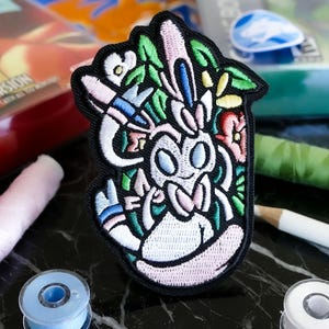 Peut inclure: Écusson brodé représentant un personnage de dessin animé avec un motif floral. Le patch a une bordure noire et présente du rose, blanc, vert, bleu et jaune. Posé sur une surface en marbre noir, avec des fournitures de couture.