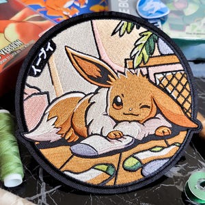 Op de afbeelding: Geborduurde patch met Eevee van Pokémon, in bruin, oranje en crème, knipogend. De ronde patch heeft een zwarte rand en Japanse tekst. Naaigarenspoelen en een Pokémon-gamebox staan op de achtergrond.