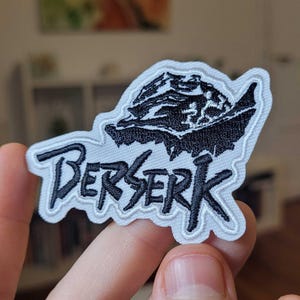 Peut inclure: Un patch brodé blanc avec des points noirs. Le patch représente une illustration en noir et blanc d'un personnage de la série d'anime Berserk, ainsi que le mot "Berserk" en lettres noires.
