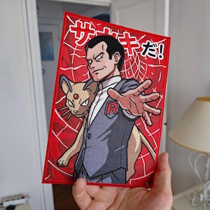 Puede incluir: Un parche rectangular bordado que muestra a un hombre con traje gris con un emblema R rojo y una criatura beige similar a un gato. El fondo es rojo con un patrón de telaraña blanco y texto japonés en la parte superior. Borde rojo y detalles cosidos.
