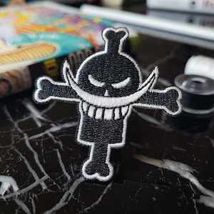 May include: Black and white embroidered patch of a skull with a crossbones and a large grin. The skull has a single eye and a mustache.