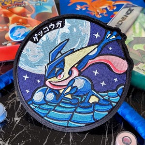 Könnte beinhalten: Ein kreisförmiger bestickter Aufnäher mit der blau-gelben Pokemon-Figur Quajutsu vor einem dunkelvioletten Sternenhimmel mit einem großen Mond. Der japanische Text "ゲッコウガ" ist oben links am Rand des Aufnähers in Weiß eingestickt.