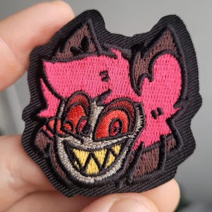 Puede incluir: Parche bordado con un personaje de dibujos animados con una cara rosa, ojos rojos y una amplia sonrisa con dientes amarillos. El parche tiene un borde negro y detalles marrones, creando un diseño detallado. Ideal para chaquetas, bolsos u otros artículos de tela.