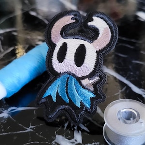Op de afbeelding: Geborduurde patch met een gestileerd personage met een wit gezicht, zwarte ogen en een blauwe cape. De patch heeft een zwarte rand. Blauw garen en een spoel garen zijn ook zichtbaar.