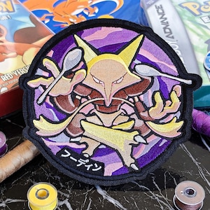 May include: A circular embroidered patch featuring a yellow and brown creature with pointed ears, holding two silver spoons. The background is a purple swirling pattern with a black border. The Japanese text "フーディン" is stitched in white at the bottom.