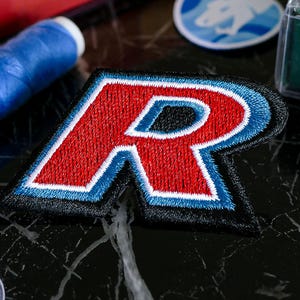 Peut inclure: Écusson brodé avec la lettre "R" rouge, un contour blanc et une bordure bleue. L'écusson est posé sur un fond noir, avec des fournitures de couture en arrière-plan. Idéal pour les vêtements ou accessoires.