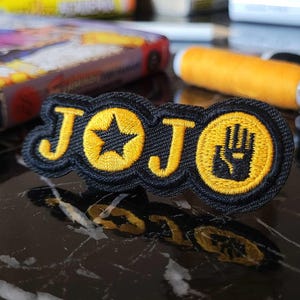 Può includere: Toppa ricamata nera e gialla con la parola "JOJO" in un font stilizzato. La toppa presenta una stella e una mano con un solo dito esteso.