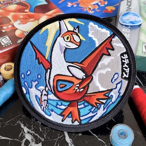 Può includere: Toppa ricamata raffigurante una creatura bianca e rossa con dettagli arancioni, su sfondo blu e bianco. La toppa ha un bordo nero e testo giapponese. Materiali da cucito e custodie di videogiochi circondano la toppa.