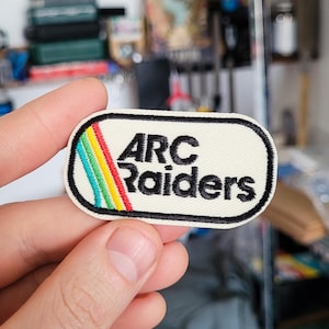 May include: Embroidered patch with the text "ARC Raiders" in black, set on a cream-colored background. A rainbow-colored stripe accents the left side. The patch has a black border.