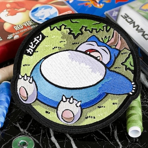 Puede incluir: Parche bordado con un personaje Snorlax durmiendo en azul, blanco y beige, rodeado de follaje verde. El parche tiene un borde negro y texto japonés. En el fondo se ven cartuchos de juego y carretes de hilo.