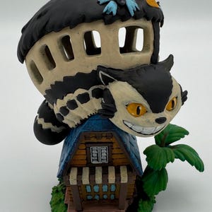 May include: A whimsical figurine featuring a grinning cat perched atop a miniature house. The cat has black and cream fur with yellow eyes. The house is beige with a black roof and blue accents. Green foliage adds a touch of nature.