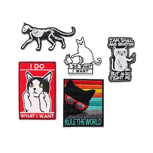 Pode incluir: Uma coleção de patches bordados com desenhos de gatos. Os patches incluem um esqueleto de gato preto e branco, um gato branco com o texto "I DO WHAT I WANT", um gato preto usando óculos de sol com o texto "RULE THE WORLD" e outros desenhos com tema de gato.
