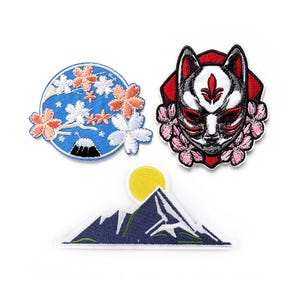 Può includere: Tre toppe ricamate da stirare con disegni di ispirazione giapponese. La prima toppa è un cerchio blu con un ramo di ciliegio bianco e una montagna nera. La seconda toppa è una maschera di volpe rossa e bianca con fiori di ciliegio rosa. La terza toppa è una montagna blu con un sole giallo.