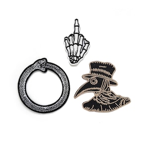 Pode incluir: Três patches bordados para passar a ferro: uma cobra preta num círculo, um médico da peste negro com um bico comprido e uma mão de esqueleto branca a fazer o dedo do meio.