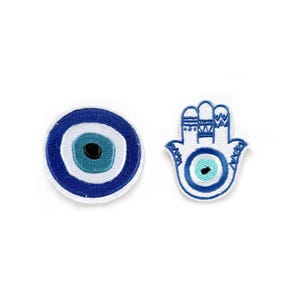 Puede incluir: Dos parches bordados, uno con un ojo azul mal de ojo y el otro con una mano de hamsa blanca con un ojo azul mal de ojo en el centro.