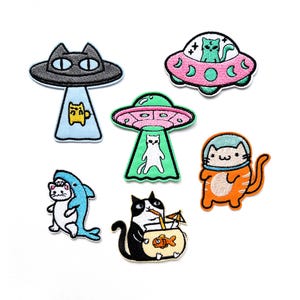 May include: Set of nine embroidered iron-on patches featuring cats in various scenes. The patches include a cat in a spaceship, a cat in a shark, a cat in a spacesuit, a cat drinking a cocktail, and a cat in a UFO. The patches are all colourful and whimsical.