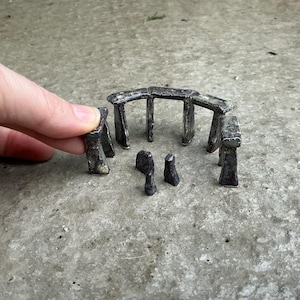 May include: Miniature Stonehenge replica in a weathered, dark gray finish. The set includes upright stones and lintels, arranged to mimic the ancient monument. The small-scale model is designed for display or as a unique decorative piece.