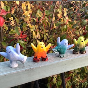 May include: Four ceramic figurines with smiling faces and outstretched arms. Each figurine is painted in a different colour: blue, orange, green, and yellow. The figurines are arranged in a row on a wooden surface.