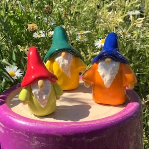 May include: Three ceramic gnomes with colourful hats and painted faces. The gnome on the left is wearing a red hat and a green shirt. The gnome in the middle is wearing a green hat and a yellow shirt. The gnome on the right is wearing a blue hat and an orange shirt.