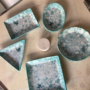Peut inclure: Ensemble de plats de service en céramique de formes variées : carrée, rectangulaire, ovale, triangulaire et ronde. Chaque pièce présente un motif de bulles turquoise et blanches et un petit motif de tortue. Une petite pièce en céramique blanche ronde avec les lettres "L & L" est également présente.