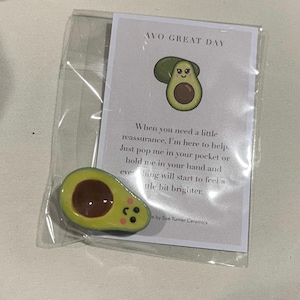 May include: A ceramic avocado charm with a smiling face, packaged with a card that reads "AVO GREAT DAY." The card includes a message about reassurance. The avocado is yellow, brown, and green.