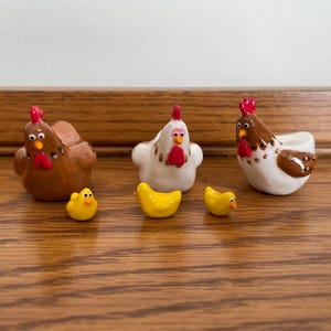 May include: A collection of ceramic chicken figurines and yellow chick figurines. The chickens are brown and white with red accents. The chicks are small and yellow. The figurines are arranged on a wooden surface.