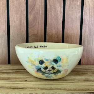 May include: A cream-coloured ceramic bowl featuring a watercolour-style illustration of a panda face surrounded by colourful paint splatters. The interior rim of the bowl displays the handwritten black text that reads black and white.