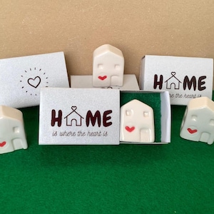 Peut inclure: Cinq petites maisons en céramique blanche avec des cœurs rouges sur le devant. Chaque maison est dans une petite boîte blanche avec une finition argentée scintillante. Les boîtes portent le mot "HOME" imprimé avec une petite icône de maison et le texte "is where the heart is".