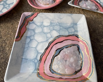 May include: A collection of decorative ceramic dishes with a geode design. The dishes feature a light blue background with a bubble pattern, pink and blue geode-like rings, gold accents, and a cluster of crystals in the center. The shapes include square, oval, and triangular.