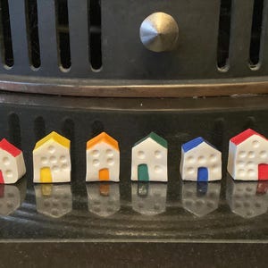 May include: Eight miniature ceramic houses in various colours, including purple, red, yellow, orange, green, blue, and white. Each house has a white base and a different coloured door and window.
