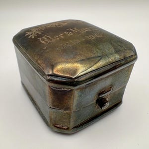 May include: A rectangular metal box with a tarnished silver finish, featuring a hinged lid with an engraved branch motif and the text "Alec to Mary 1913-1964". The box has a small, centred push-button latch on the front side for opening.