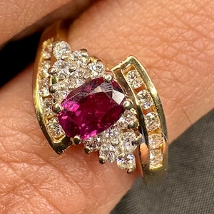 Op de afbeelding: Een gouden ring met een centrale ovaal geslepen donkerroze edelsteen, omringd door een cluster van kleine ronde heldere stenen. De ring heeft een bypass-ontwerp met extra heldere stenen in kanaalzetting langs de zijkanten van de gouden band.
