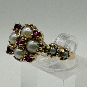 May include: A gold ring featuring a cluster of round white pearls and small red gemstones arranged in a floral pattern. The band extends along the finger with two additional small pearls set in gold, creating a detailed and ornate jewellery piece.