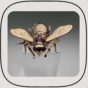 Puede incluir: Broche de abeja dorada con detalles de rubíes. La abeja tiene alas detalladas y un cuerpo texturizado. El broche está sobre una superficie transparente, mostrando su intrincado diseño y artesanía. Una pieza de joyería clásica.