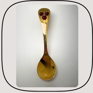 Könnte beinhalten: Ein goldfarbener Löffel mit einem Kirschdesign am Griff. Die Kirschen sind rot mit grünen Blättern. Der Löffel hat eine abgerundete Schüssel und einen langen, schlanken Griff. Der Löffel wird wahrscheinlich zum Servieren oder Essen verwendet.