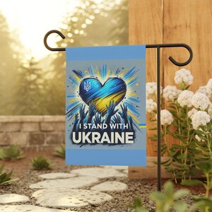 May include: A blue and yellow heart with the Ukrainian flag and the text "I Stand With Ukraine" on a grey background. The heart is surrounded by raised hands.