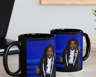 Puede incluir: Dos tazas de café negras con una fotografía de una mujer con blazer negro y camisa blanca. La mujer señala con ambas manos. El fondo es azul. Las tazas están sobre una superficie clara.