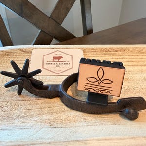 May include: A dark brown metal spur featuring a rowel with eight points and a rectangular tan leather attachment embossed with a black floral-inspired design. A white rectangular card with the text DOUBLE K LEATHER CO. is positioned behind the spur.