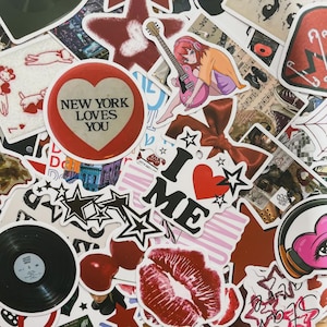 Può includere: Una collezione di adesivi colorati con vari disegni. Include un adesivo con la scritta "NEW YORK LOVES YOU" a forma di cuore, un disco in vinile e un adesivo di una ragazza che suona una chitarra rosa.