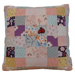 May include: A patchwork cushion with a pink background and various colourful patterns, including floral, animal print, and geometric designs. A small red ladybug is embroidered on the cushion.