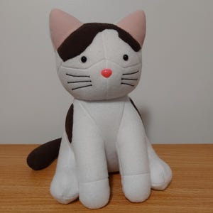May include: A white and brown plush cat toy with pink ears and a pink nose. The cat is sitting with its tail curled behind it.