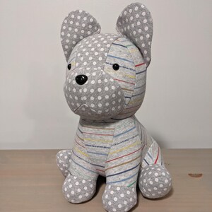 May include: A soft toy dog in a sitting pose. The head and paws are grey with white polka dots. The body is grey with multi-coloured horizontal stripes. The dog has black button eyes and a black nose.