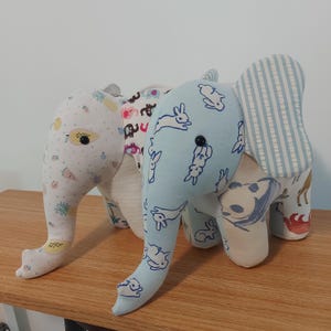 May include: A patchwork elephant soft toy with various patterned fabrics. The elephant has a light blue body with white rabbit illustrations, a striped ear, and a trunk with a fruit pattern. It sits on a wooden surface.