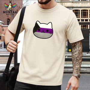 May include: A cream-coloured t-shirt with a Demisexual cat face design. The cat face has a black and white head, a purple stripe, and a grey bottom. The shirt has short sleeves and a crew neck. The brand logo "MONZAA ART LAB" is visible in the upper left corner.