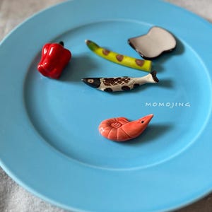 May include: A light blue ceramic plate with several small, decorative food-shaped items. These include a red pepper, a yellow and green banana, a fish, a shrimp, and a white and gray shape. The word "MOMOJING" is printed on the plate.