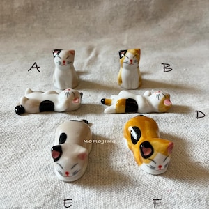 May include: Six ceramic cat figurines in different poses. The cats are white, orange, and black, with detailed faces. Some are upright, others are lying down. The figurines are small, likely a few centimetres in height.
