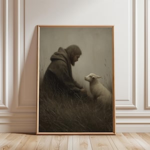 Puede incluir: Una impresión enmarcada en tonos sepia representa a una persona con una túnica con capucha interactuando con un cordero en un campo cubierto de hierba. La obra de arte tiene una calidad suave y pictórica, con tonos apagados y un enfoque en las figuras. El marco es de color madera clara.