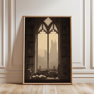 Op de afbeelding: Een ingelijste sepia-kleurige prent toont een bibliotheekinterieur met een groot boogvenster met uitzicht op een kasteel. Boekenkasten bekleden de muren, en stapels boeken en kaarsen staan op een tafel. Het kunstwerk heeft een vintage, gotische esthetiek.